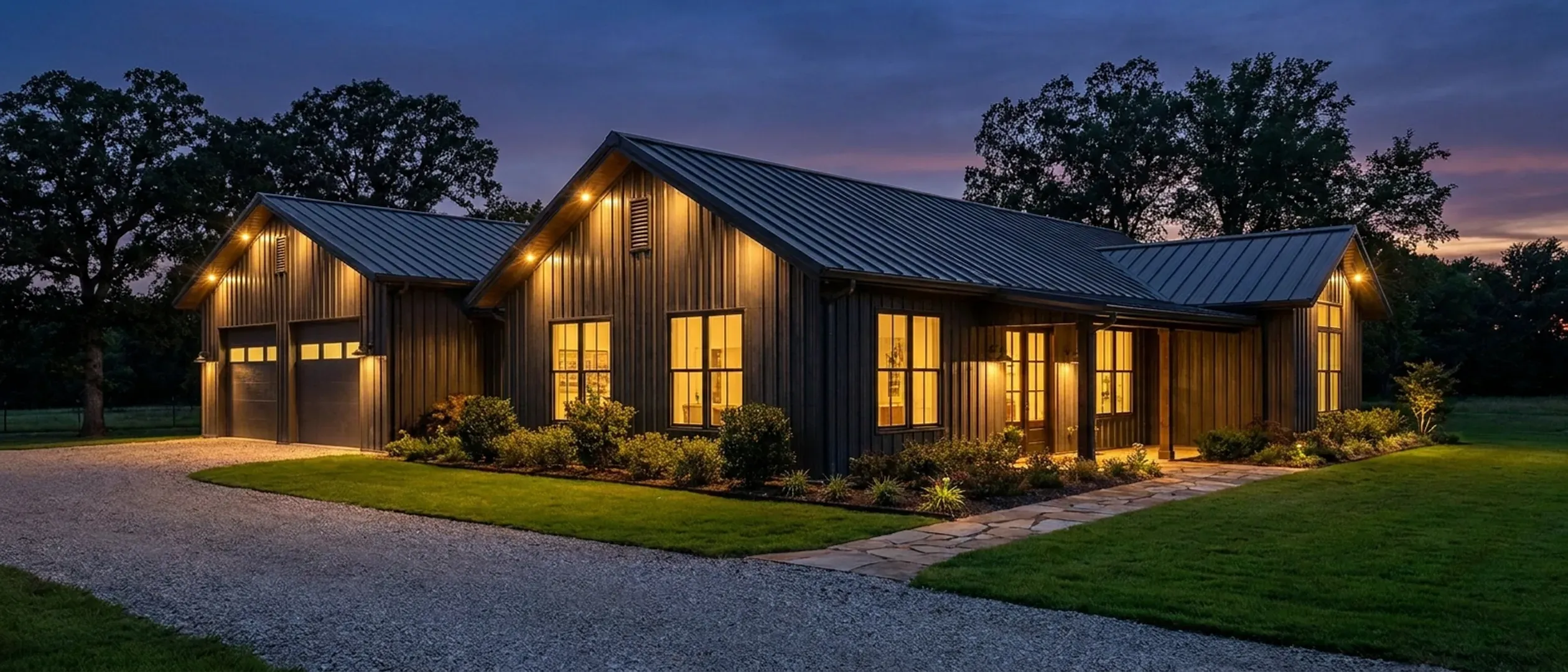 A barndominium at dusk with warm light glowing from every window and exterior lighting along the porch and roofline against a deep blue twilight sky.