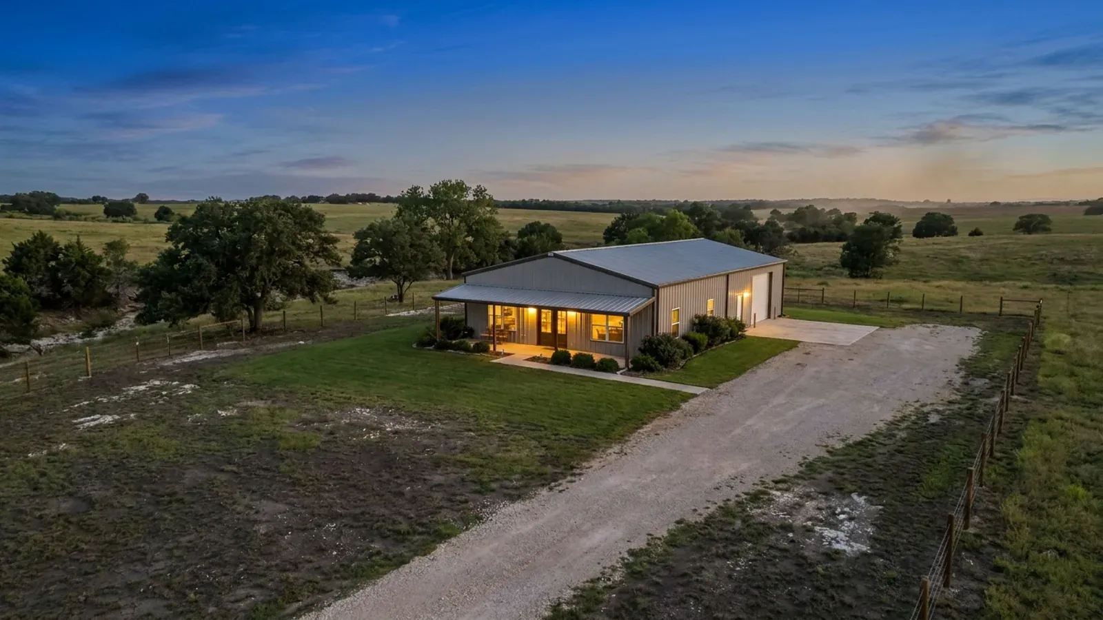 A finished barndominium at dusk with warm interior lighting glowing through its large windows.
