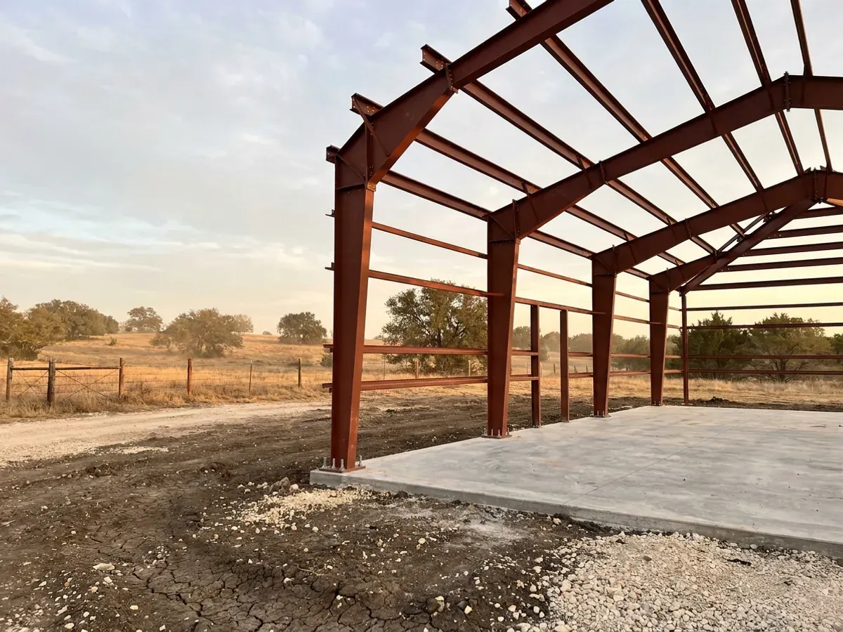 A red oxide primed structural steel frame partly erected on a concrete slab, showing bolted knee joints and anchor bolts.